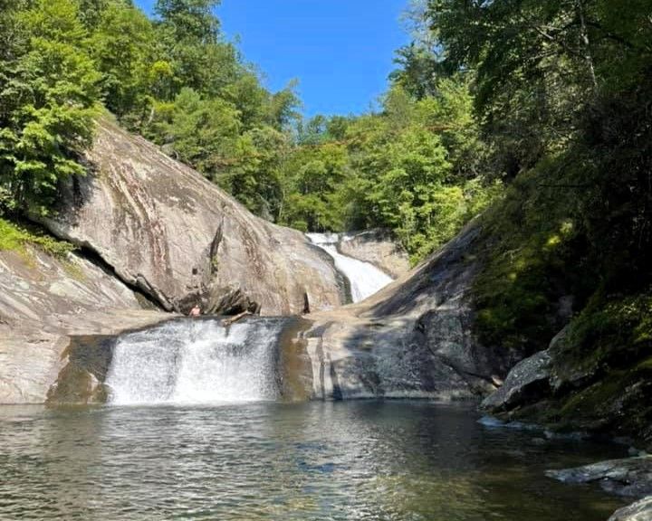 Waterfall cascading over rocky ledges into a clear pool, surrounded by green trees under a blue sky.
