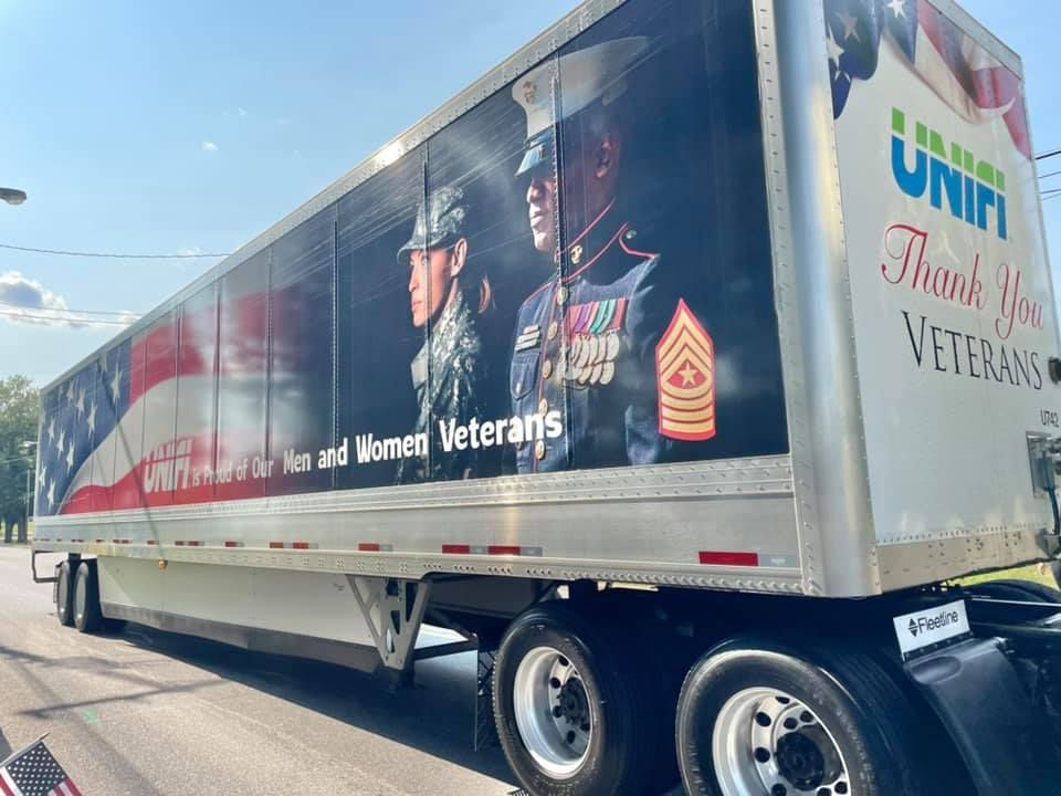 Semi-truck trailer with veterans-themed mural and Unifi logo parked on a road under a blue sky