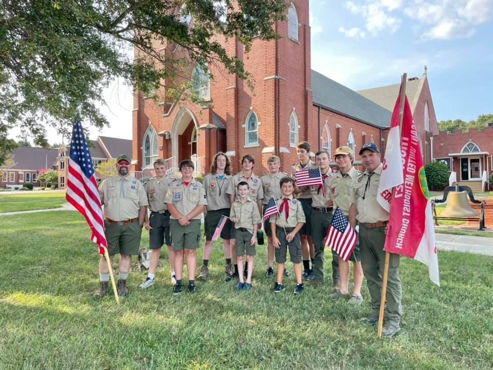 Scouts posing on a church lawn with American and red troop flags in front of a brick church.