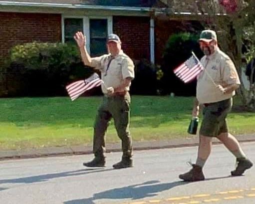 Two people walk along a street waving small American flags in front of a house.