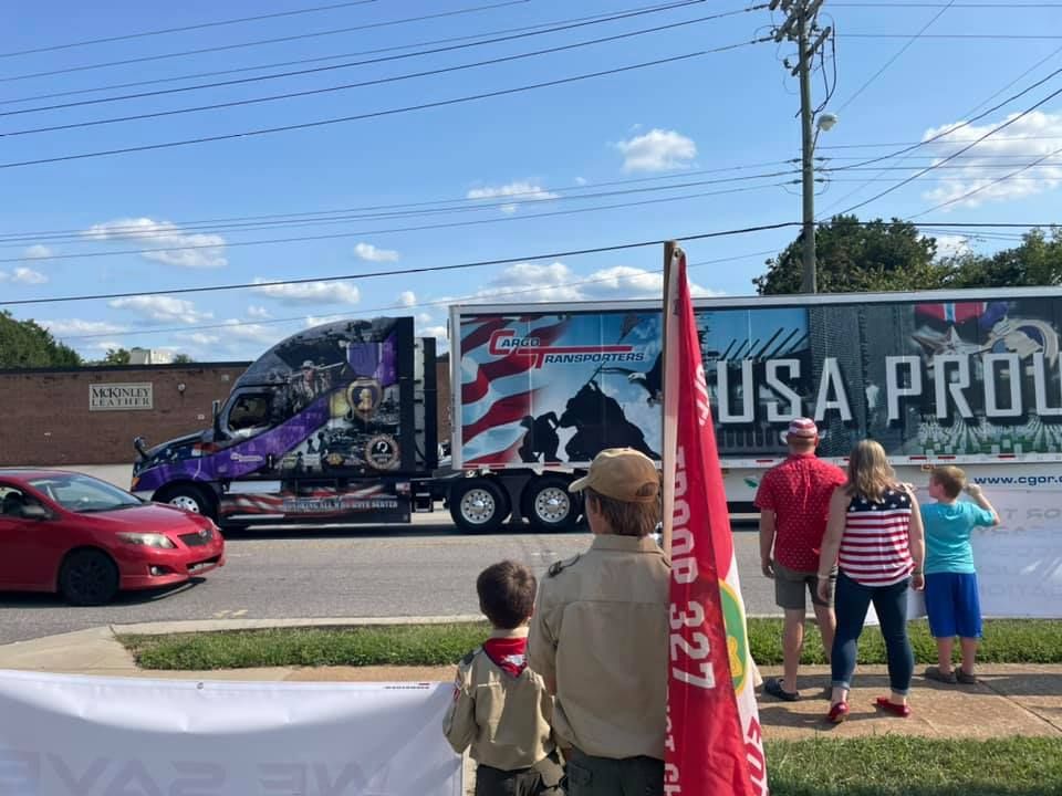 Truck convoy on a street, with spectators watching from the sidewalk under a blue sky.