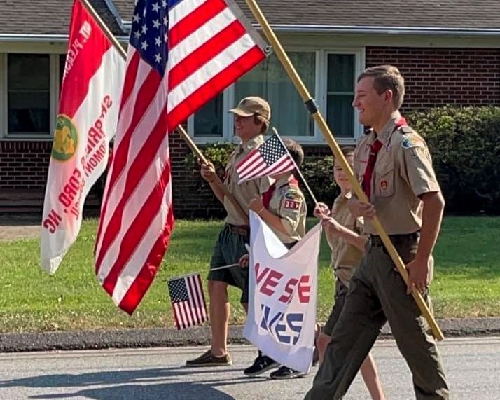 Scouts in tan uniforms carrying American and scouting flags during a parade outdoors