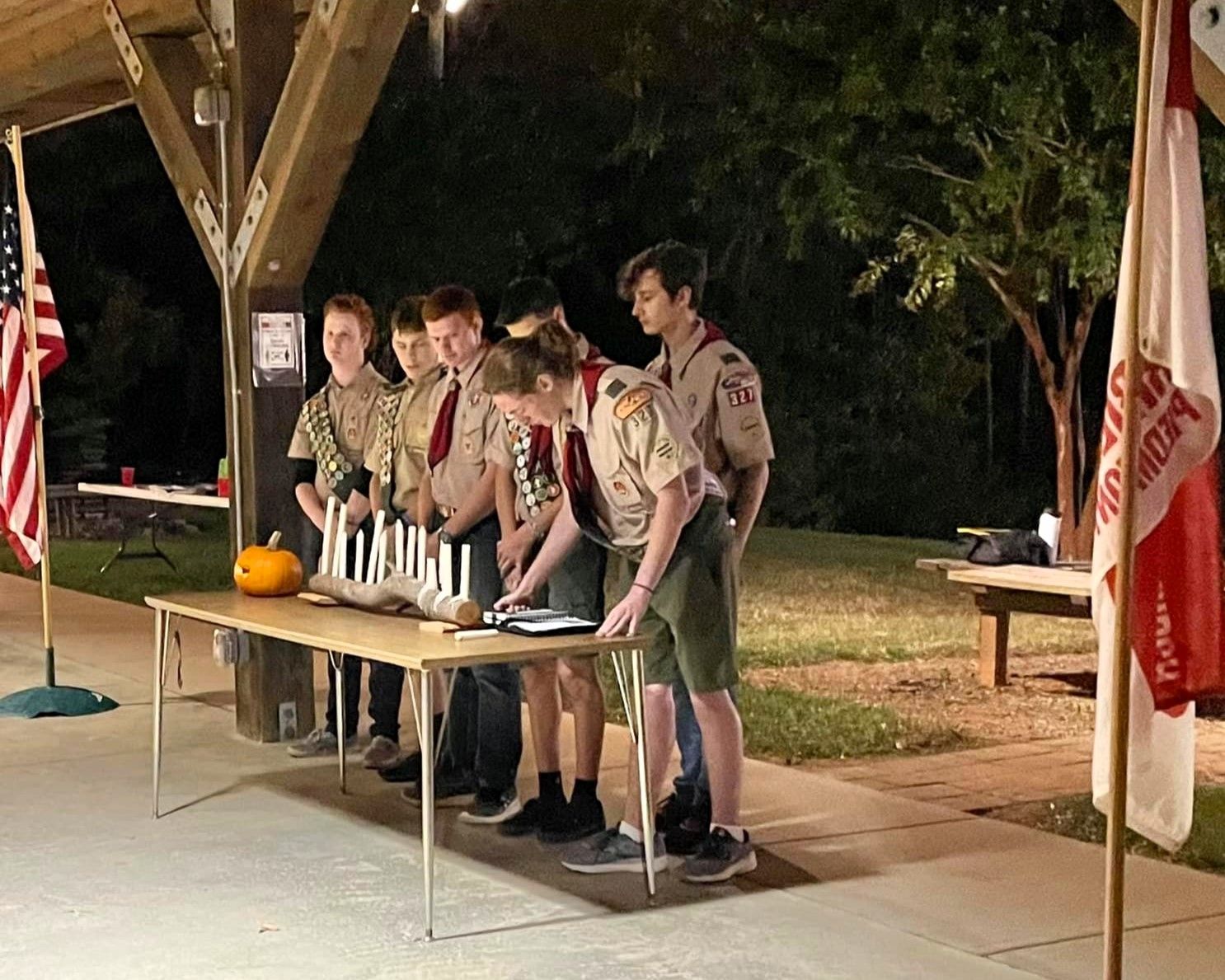 Five uniformed scouts stand behind a table with candles, under a pavilion at night.