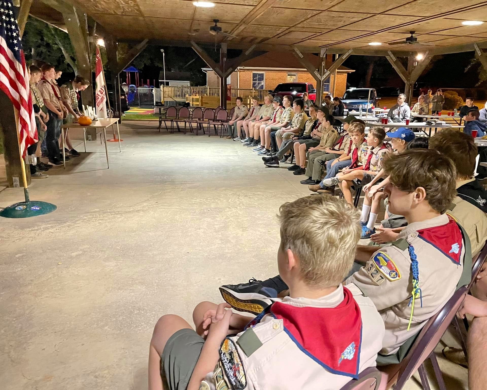 Children seated in a camp hall watching a speaker near a flag and stage area