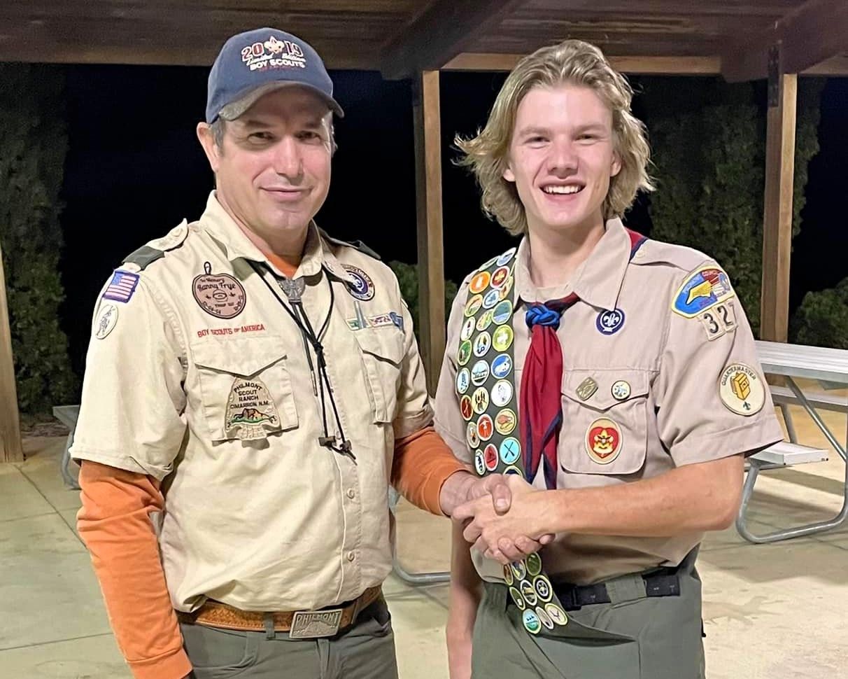 Two scouts in tan uniforms shake hands, smiling, with merit badges visible at a campsite.
