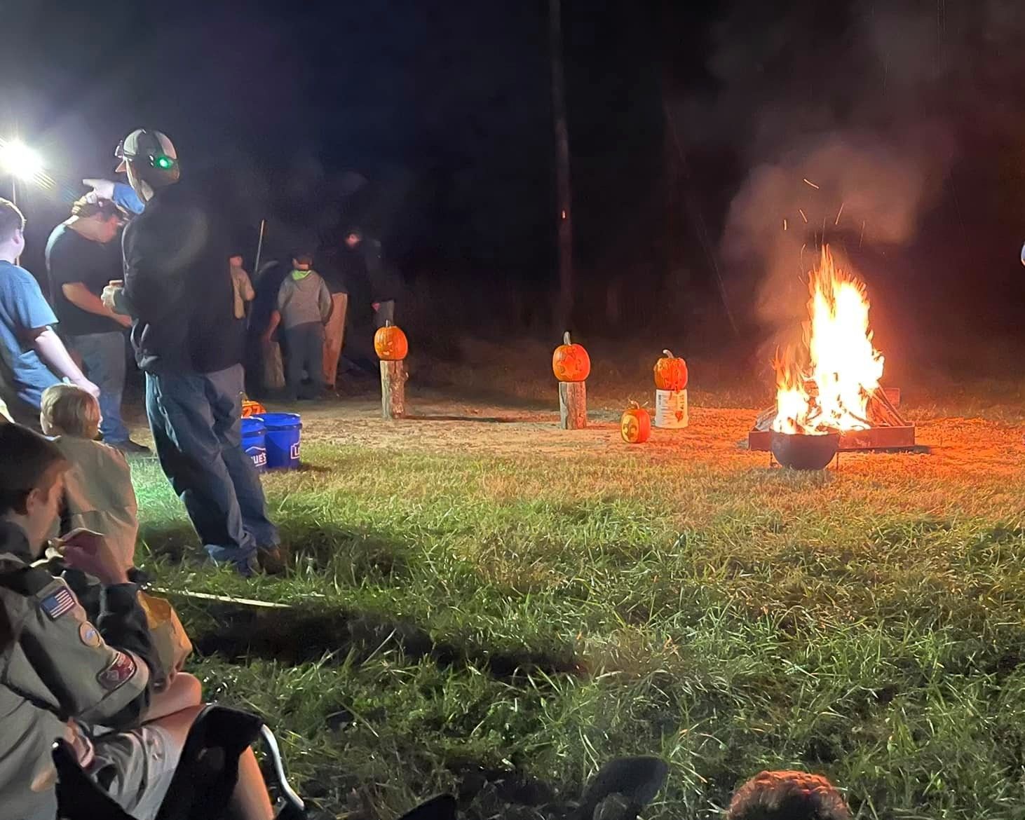 People watching a nighttime bonfire in a grassy field, with several orange-clad figures nearby.