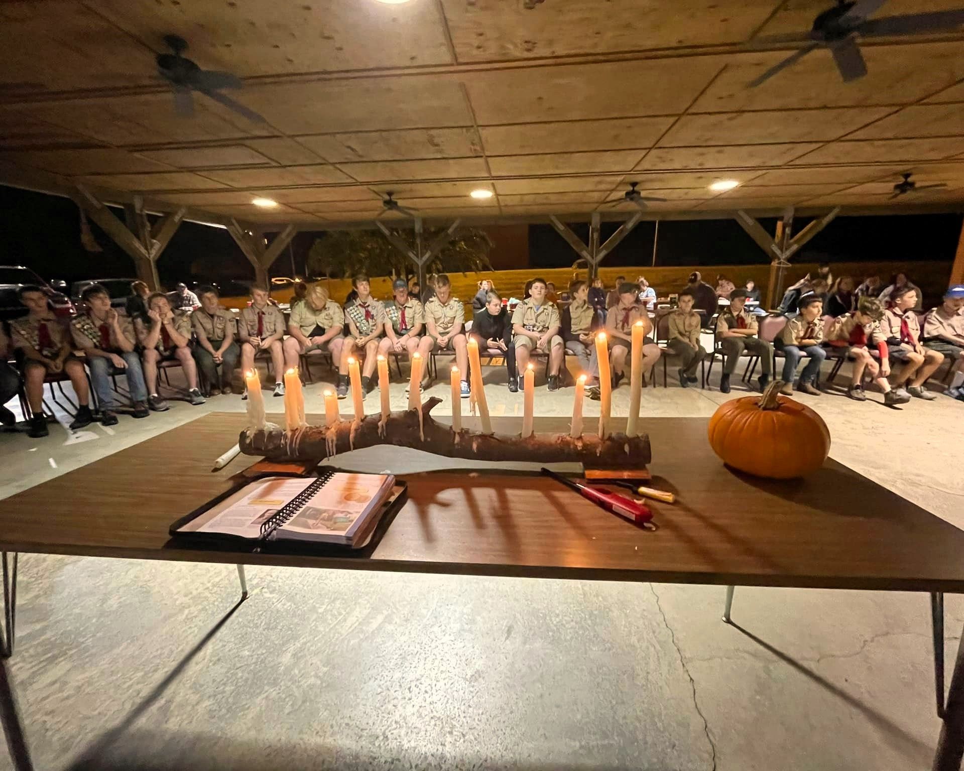 Children seated behind a candle-lit table with books and a pumpkin in a covered pavilion at night