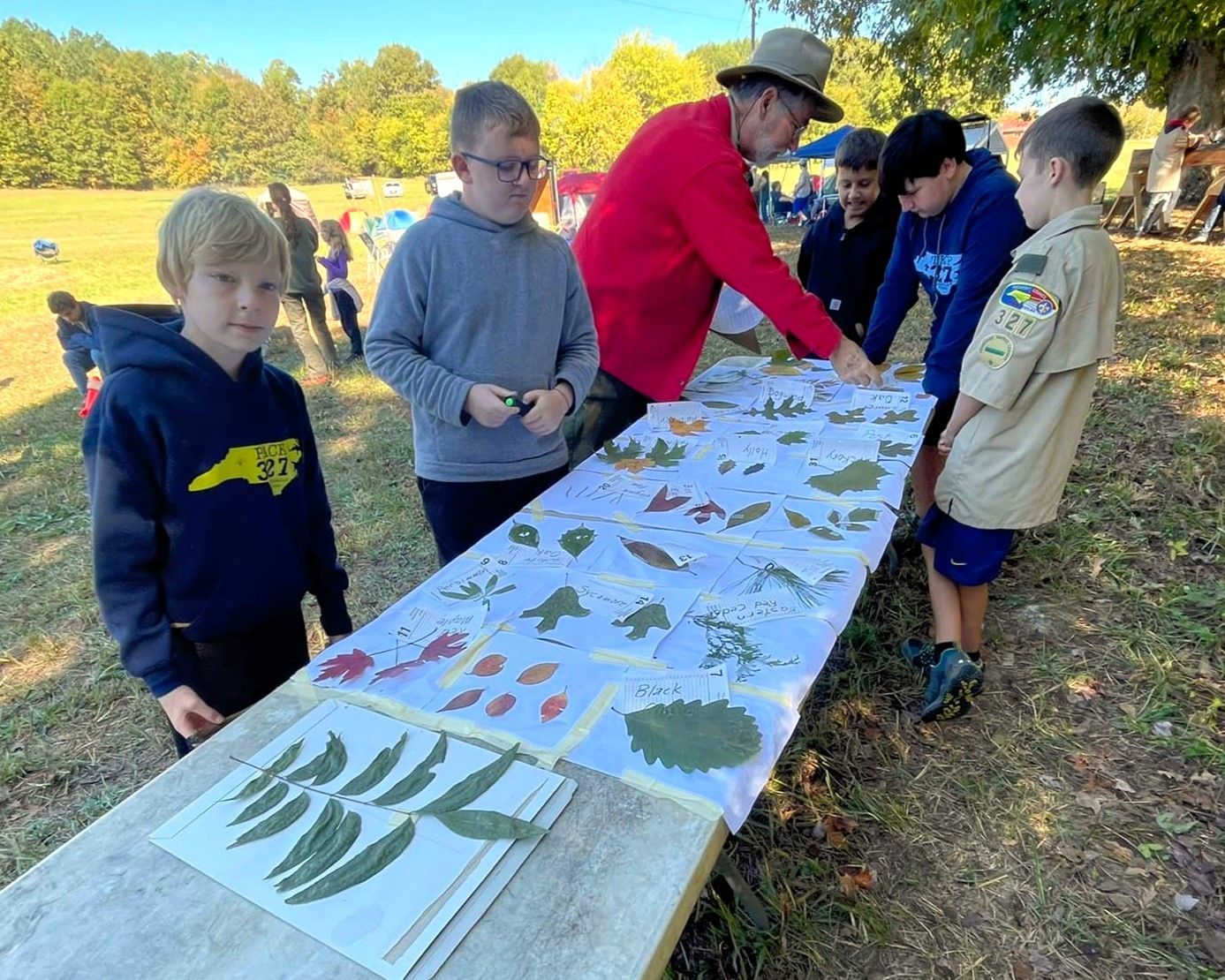 People outdoors studying leaves on a long display table with an instructor in a red jacket and hat
