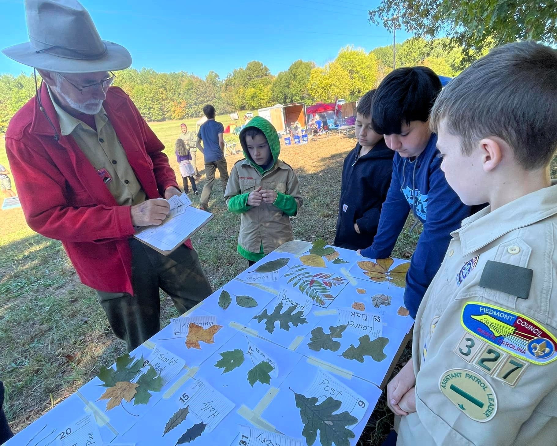 People outdoors gathering around a table with maps, listening to a guide in a hat and red jacket.