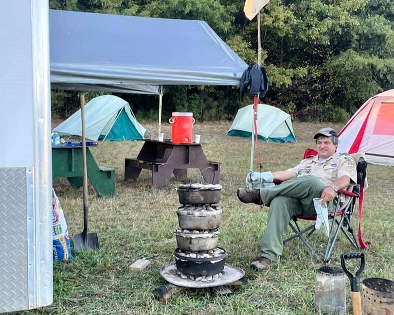 Camper relaxing beside a campfire setup under tents, with cooking gear and a red cooler nearby.