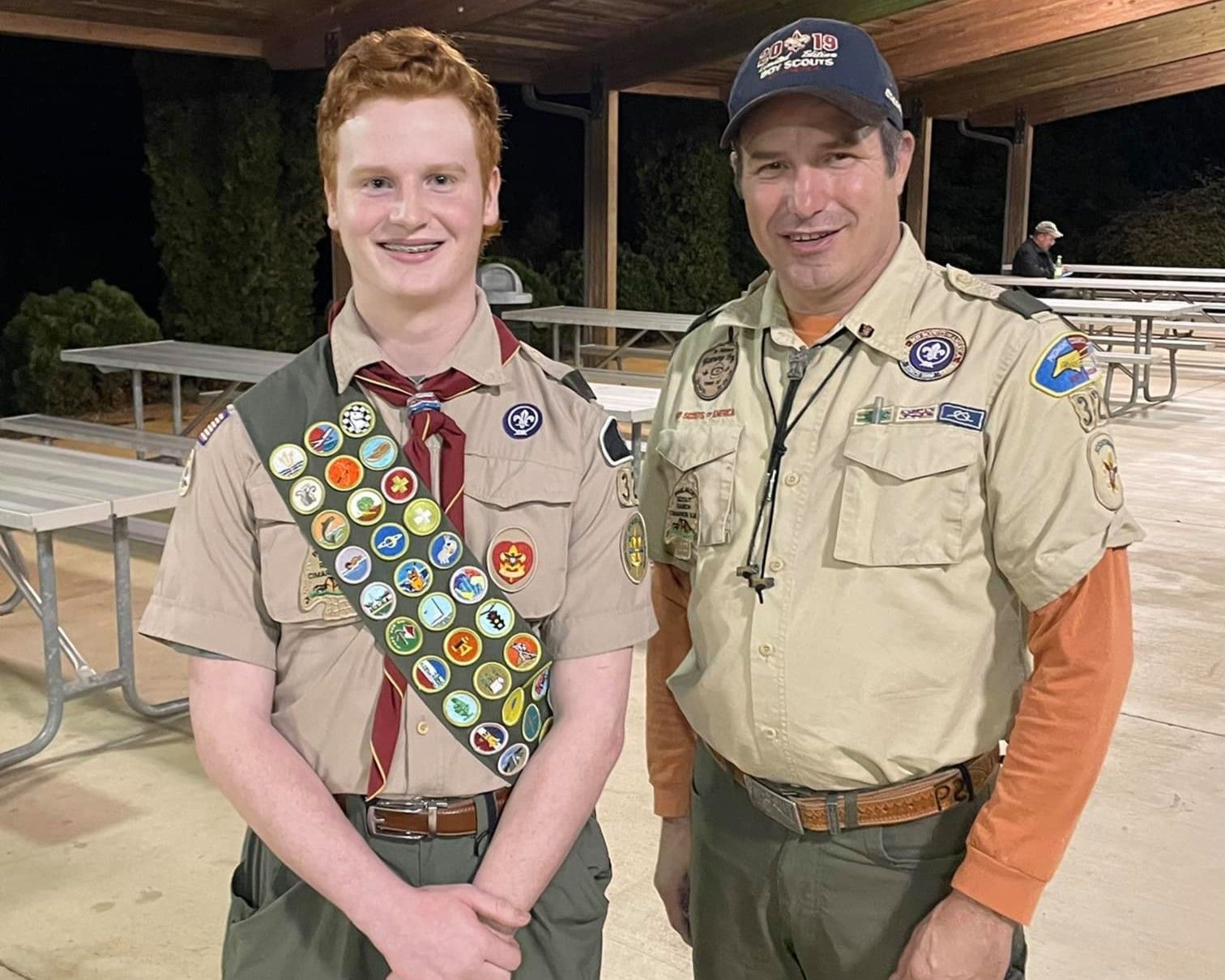 Two Scouts in tan uniforms standing together outdoors at night, smiling beside picnic tables.