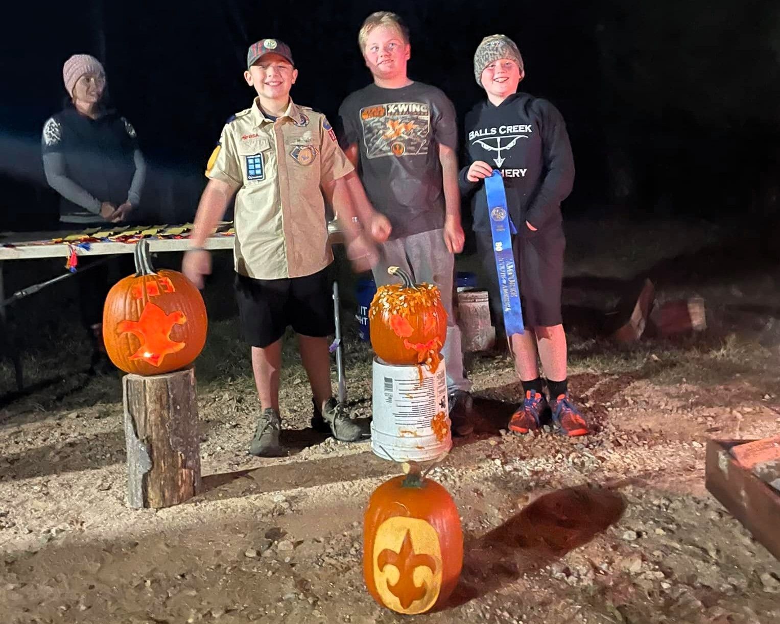 Three people stand beside carved jack-o'-lanterns at night, with a large silhouetted figure in the background.
