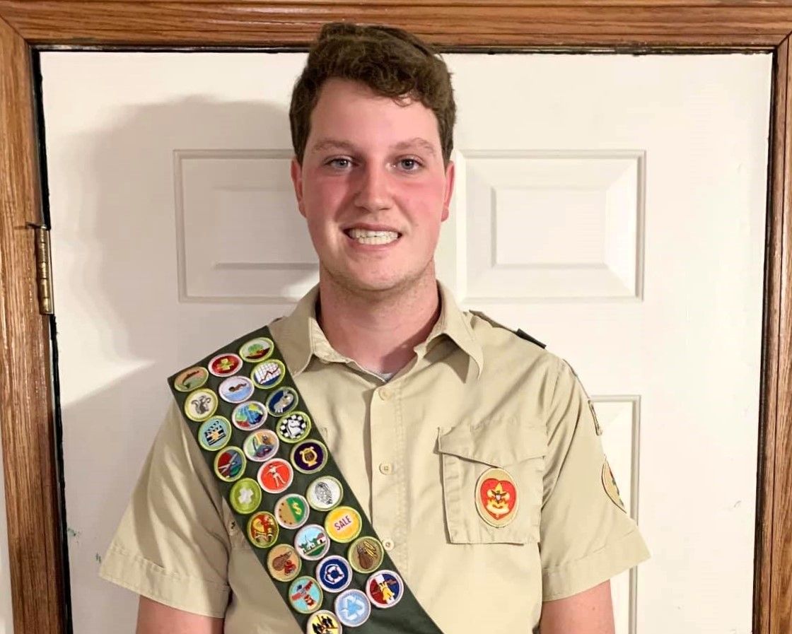 Person wearing a tan scout shirt and merit badge sash stands smiling in front of a white door.