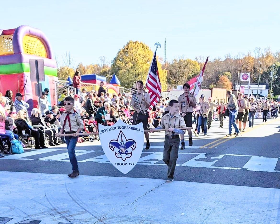 Parade marchers carrying a troop banner and U.S. flag on a sunny street, with spectators watching nearby