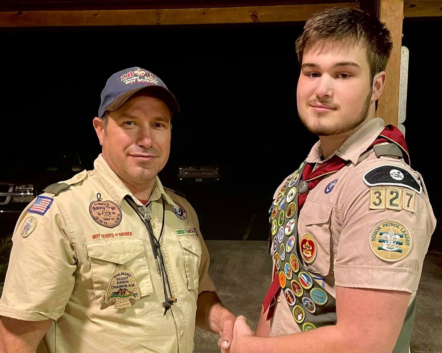 Two scouts in tan uniforms shake hands indoors, both wearing many badges and smiling at the camera.