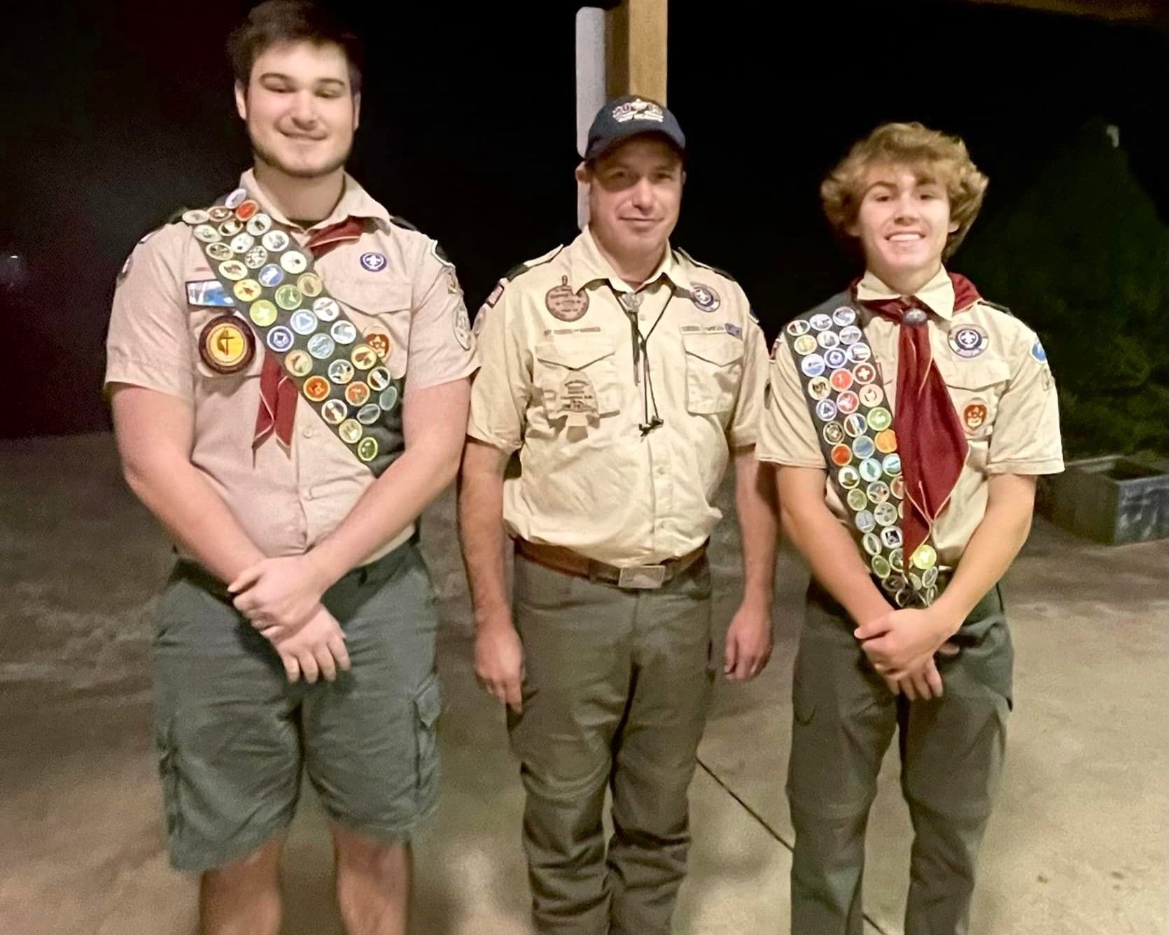 Three scouts in uniform standing outdoors at night, smiling at the camera.