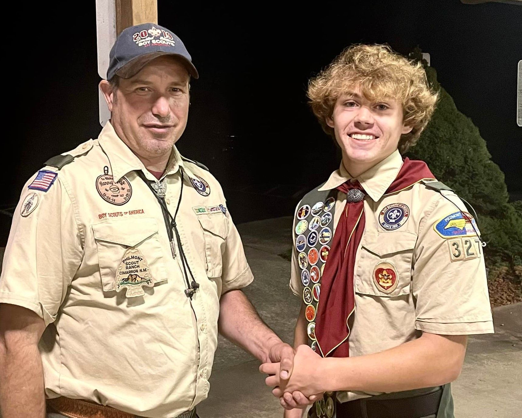 Two Scouts in tan uniforms smiling and shaking hands outdoors at night