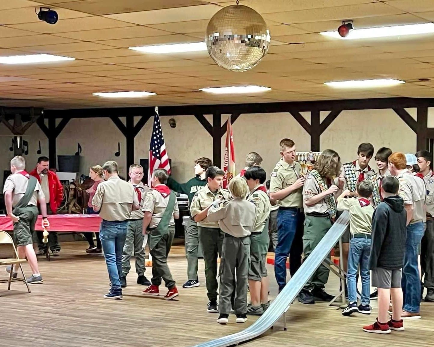 Children and adults gathered in a hall for a group activity near a red table and a slide under a disco ball.