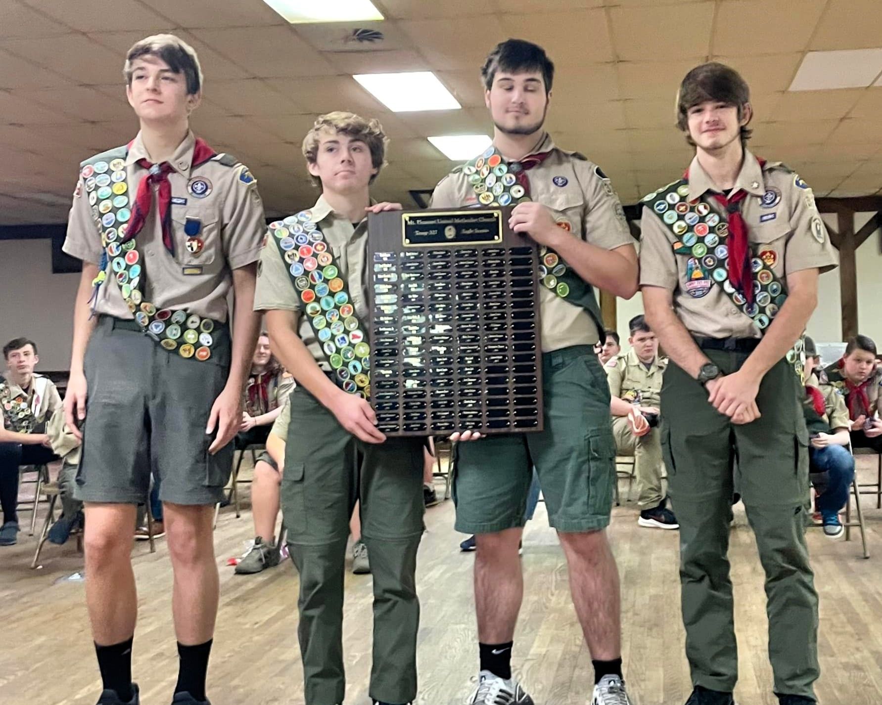 Four Scouts in uniform stand indoors, holding a large award plaque and smiling.