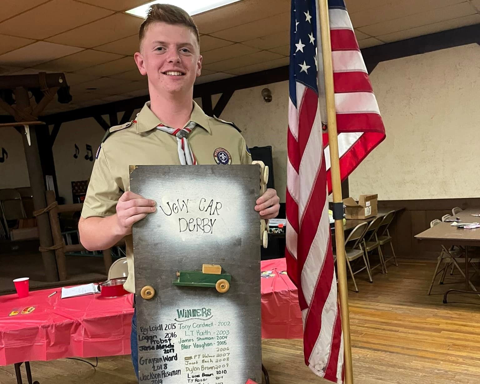 Scouts holding a metal award plaque beside a U.S. flag in a lodge hall