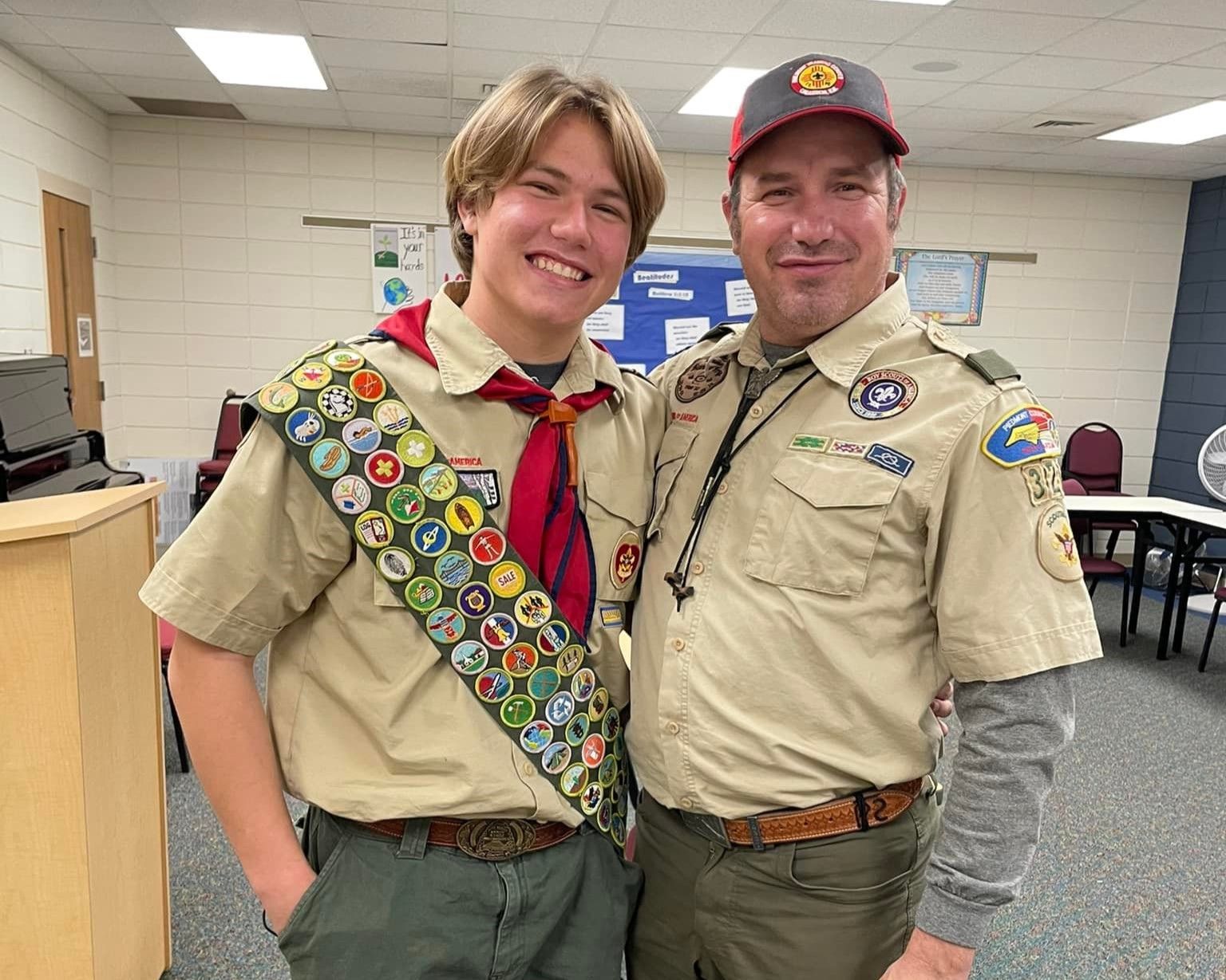 Two Scouts in tan uniforms smiling indoors, one wearing a sash of merit badges.