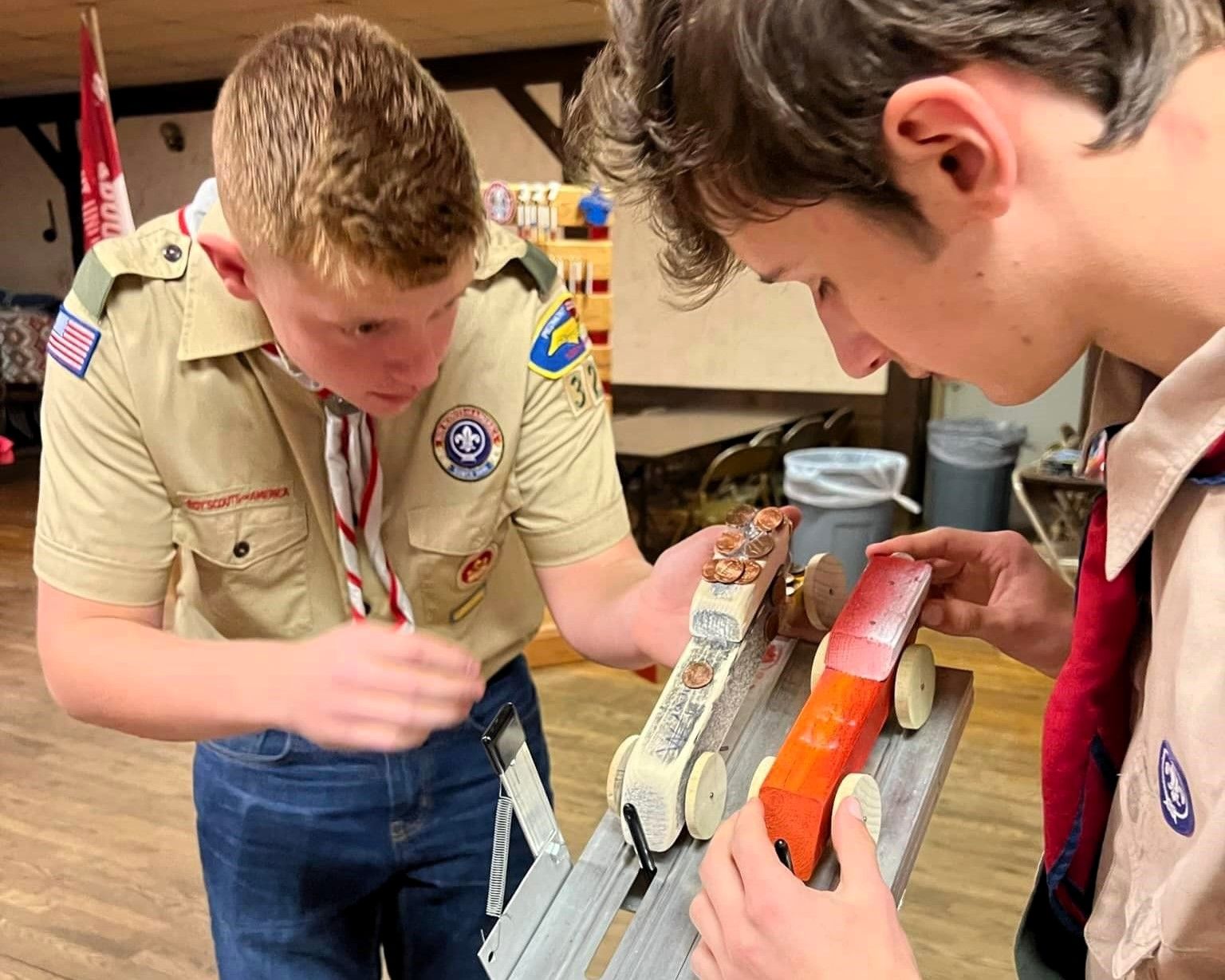 Two scouts examining a handmade launch ramp with orange and silver parts in a workshop.