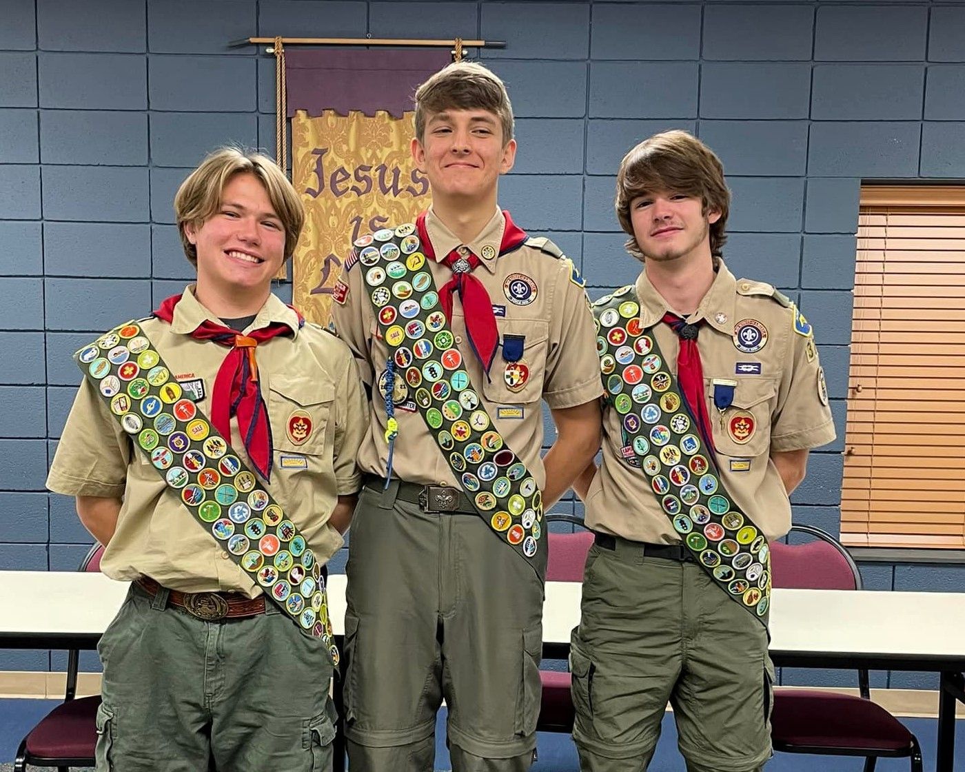 Three scouts in tan uniforms with merit badges standing together indoors in front of a blue wall.