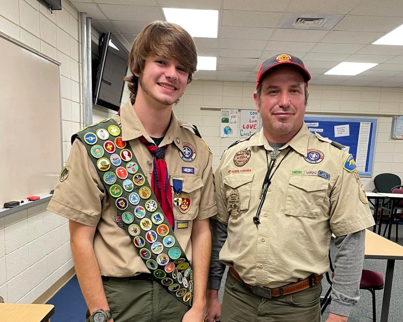Two scouts in tan uniforms standing indoors, one with merit badges and both smiling at the camera