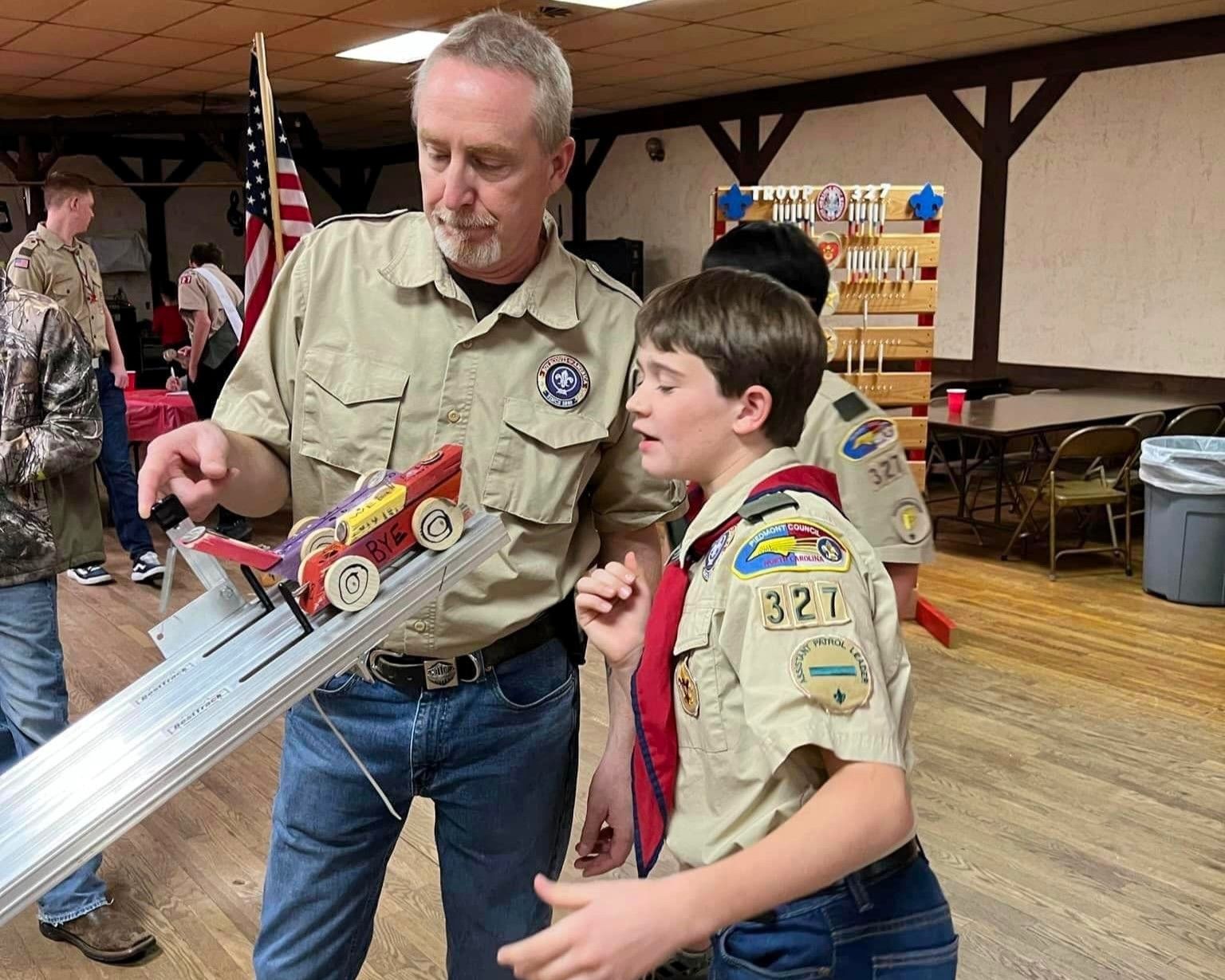 Scouts at a pinewood derby table, one adult guiding a child’s red car down a track.