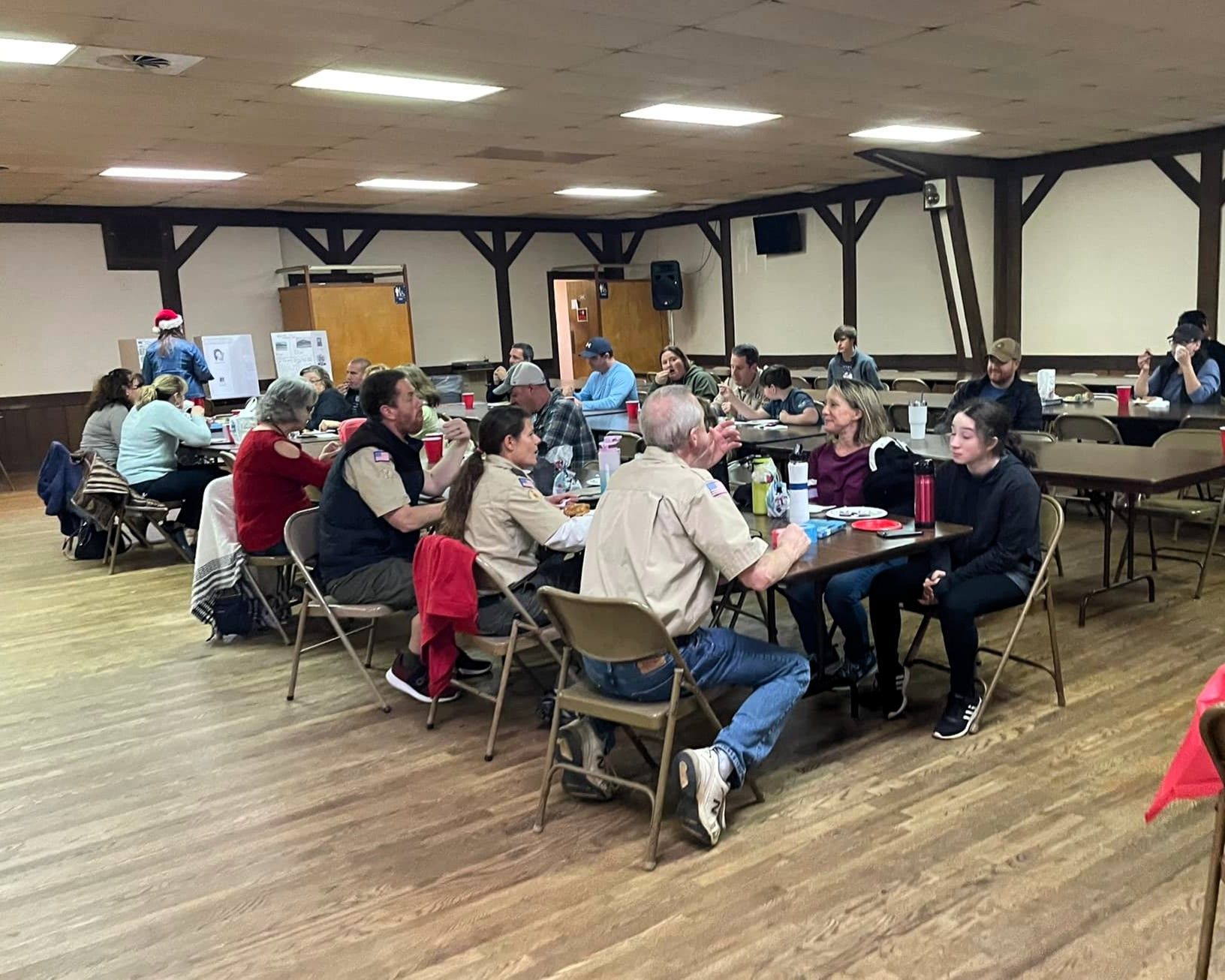 People seated at round tables in a large hall, chatting during a community meal or gathering.