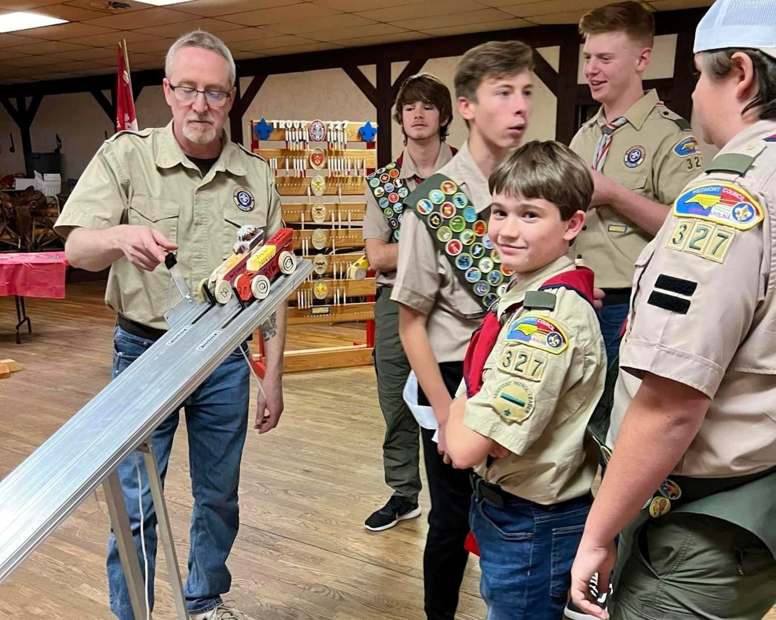 Scouts in beige uniforms gather around a tabletop race track, one boy smiling at the camera.