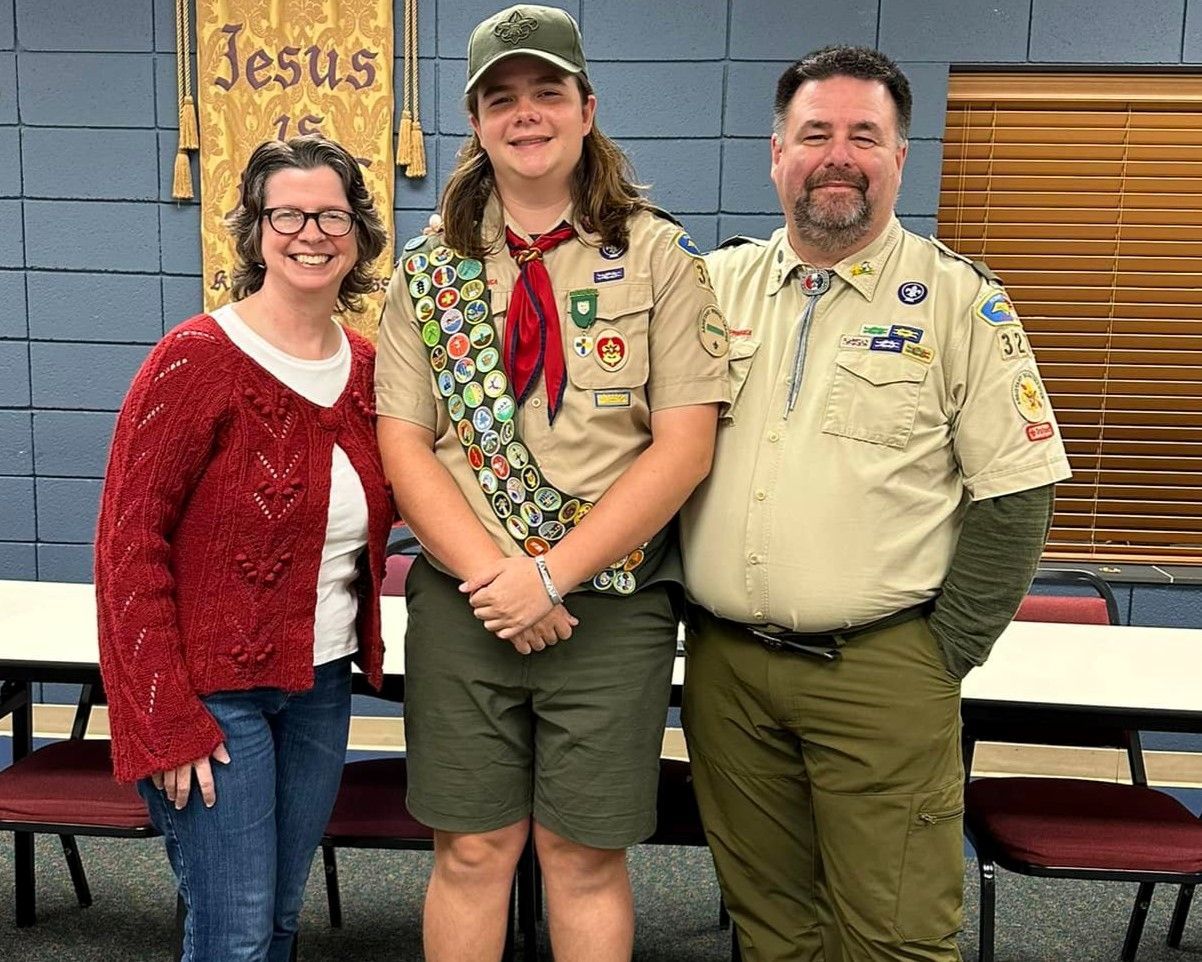Three people in scout uniforms posing indoors in front of tables and a blue wall.