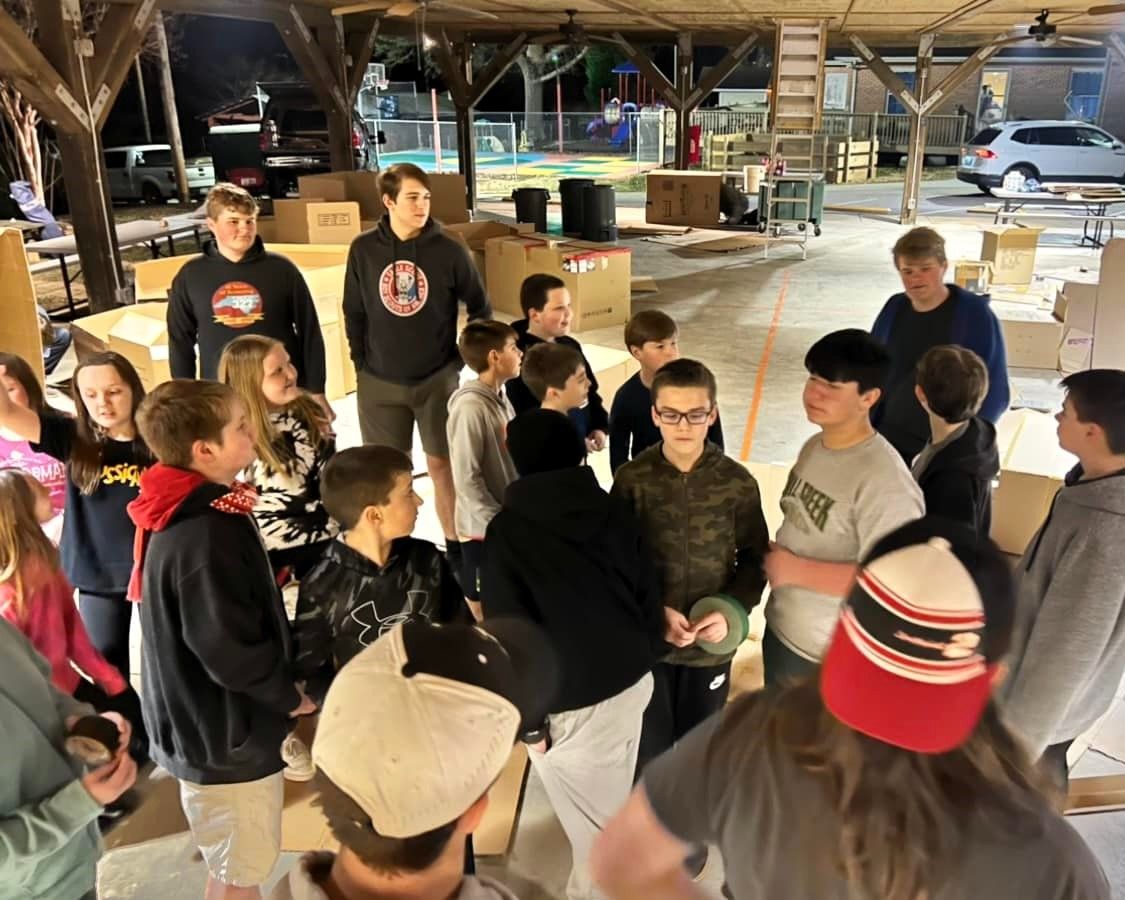 Group of people gathered in a wooden pavilion, listening to a speaker in the center.