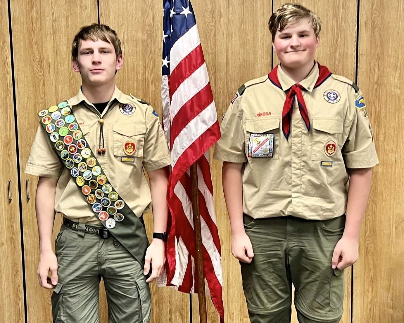 Two Boy Scouts in tan uniforms stand beside an American flag indoors.