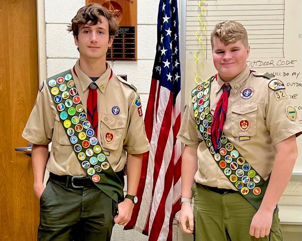 Two Scouts in tan uniforms with merit sashes stand beside an American flag indoors.