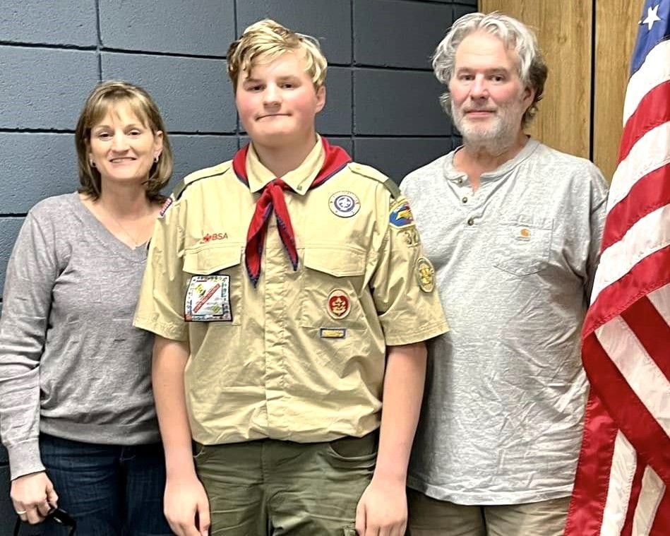 Three people standing indoors; center person in a tan scout uniform with red neckerchief, U.S. flag at right.