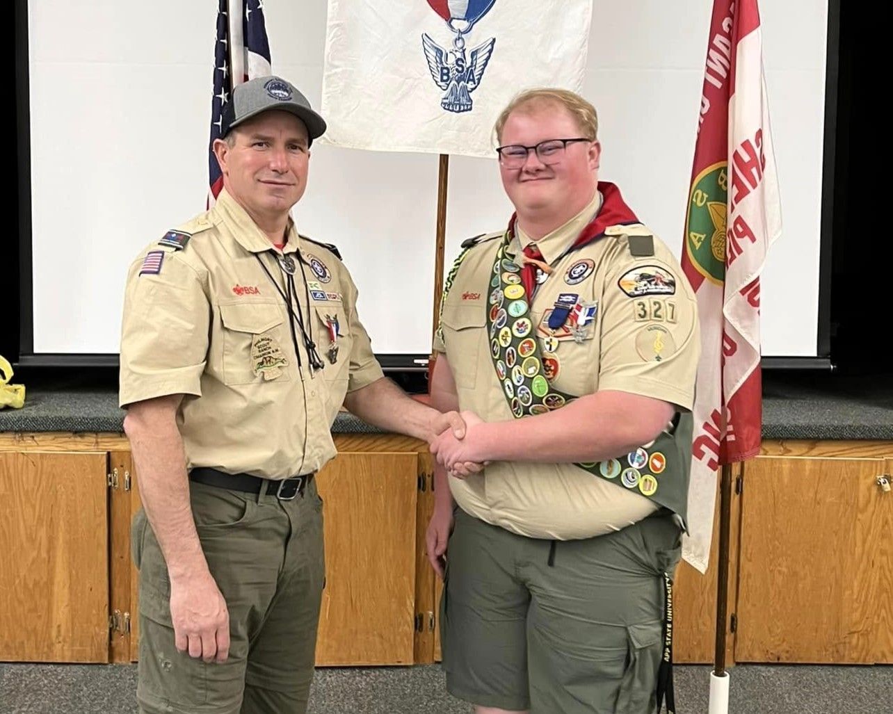 Two Scouts in tan uniforms shake hands on a stage, with flags and a presentation screen behind them.