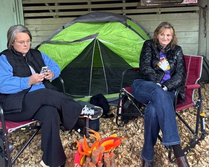 Two people seated by a green tent near a small campfire, one looking at a phone.