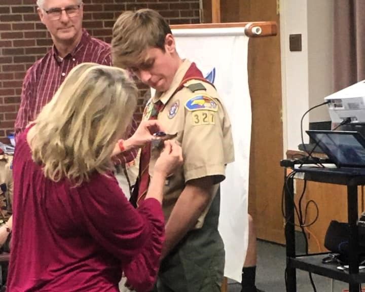 Woman pins a badge on a scout in uniform at an indoor ceremony, with a man standing nearby.