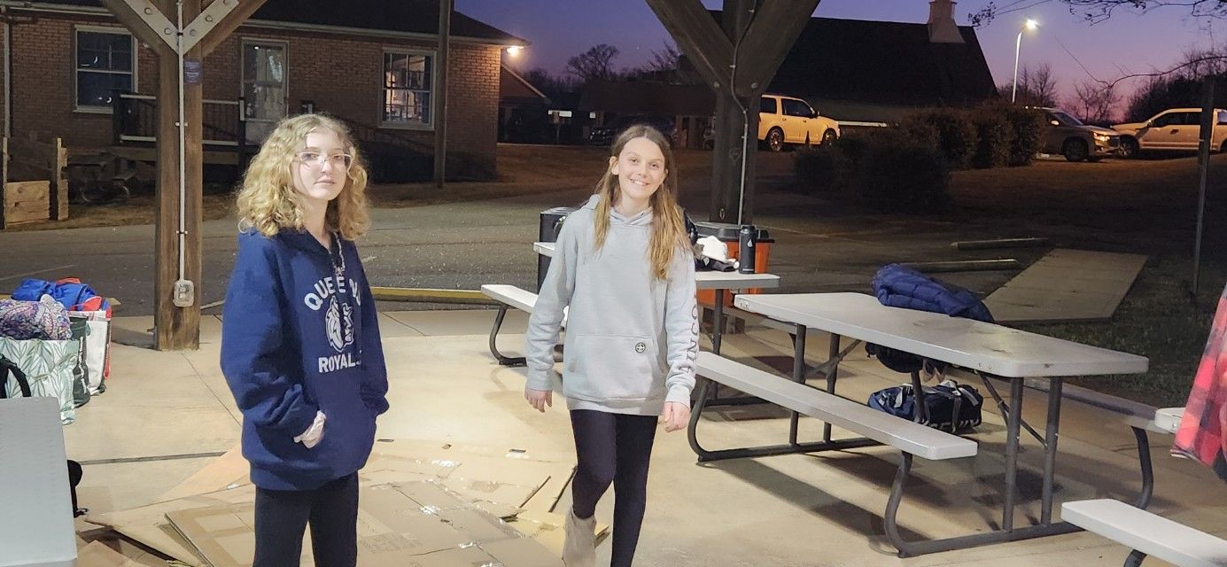 Two people standing by picnic tables outdoors at dusk in a courtyard near brick buildings