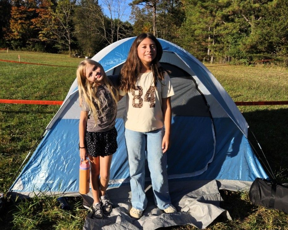 Two people standing by a blue tent in a sunny grassy campsite.