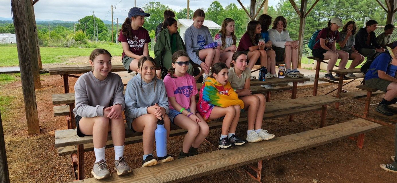 Group of children sitting on wooden bleachers outdoors under a pavilion, some smiling at the camera