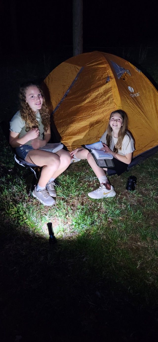 Two people sit in front of an orange tent at night, lit by a flashlight on grass.