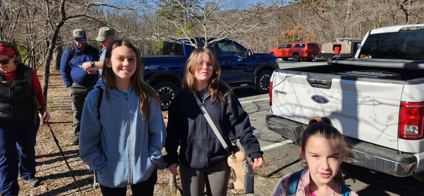 Three people smiling outdoors beside parked SUVs on a sunny day