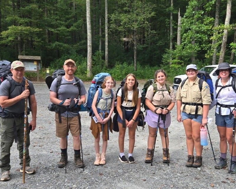 Six hikers with backpacks and walking sticks standing on a gravel trail in a wooded area