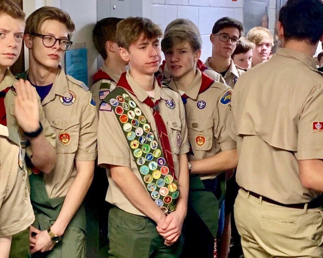 Boy Scouts in tan uniforms stand in a line indoors, one wearing a sash covered in merit badges.