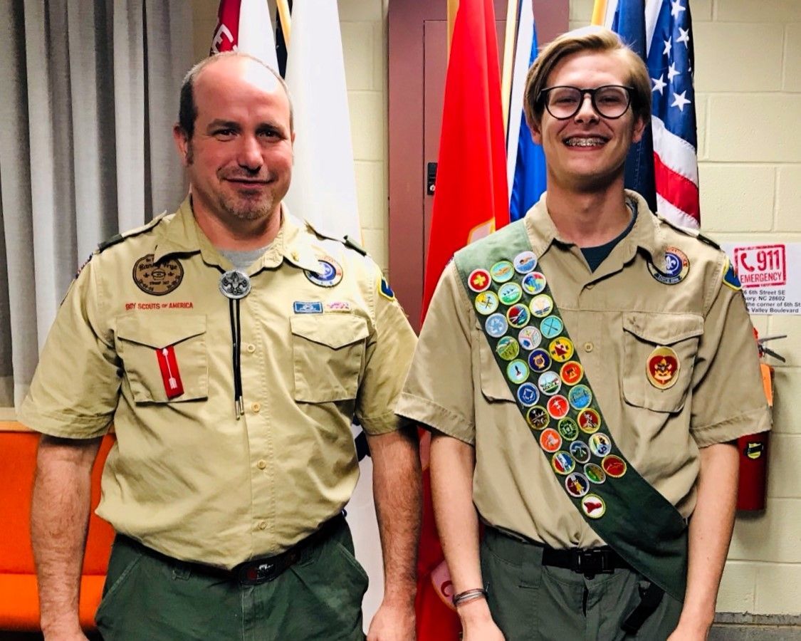 Two scouts in tan uniforms standing indoors, one with merit badges and an American flag behind them.