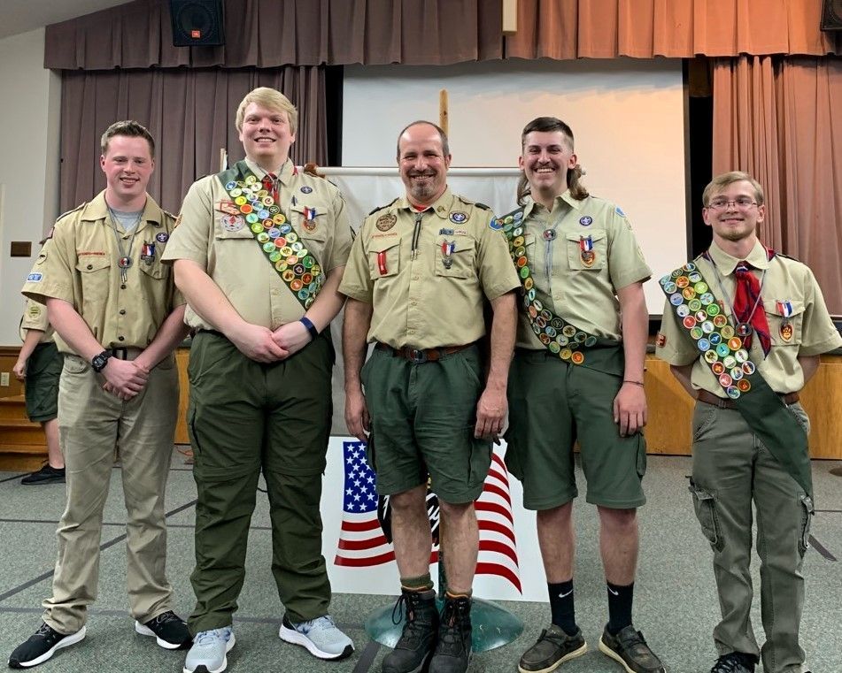 Five Scouts in khaki uniforms and sashes pose on a stage in front of an American flag backdrop.