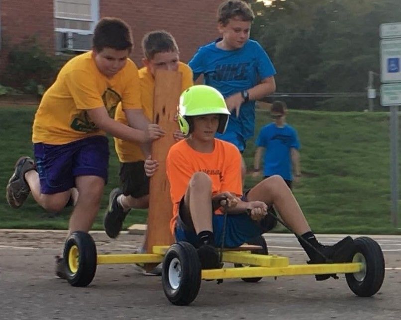 Children riding a yellow four-wheeled cart outdoors, with one wearing a neon helmet