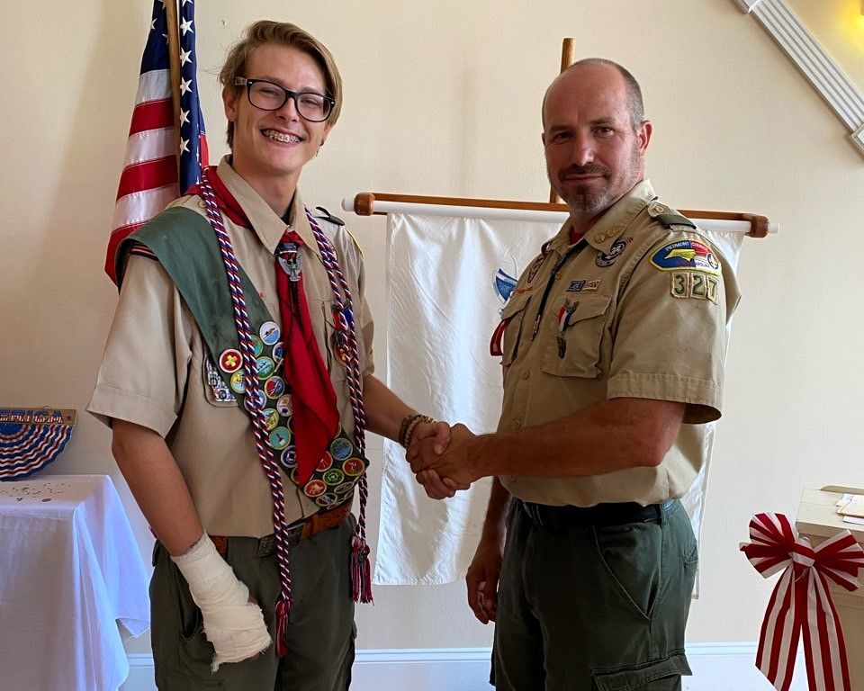 Two Boy Scouts in uniform shaking hands at a ceremony indoors, with an American flag in the background.